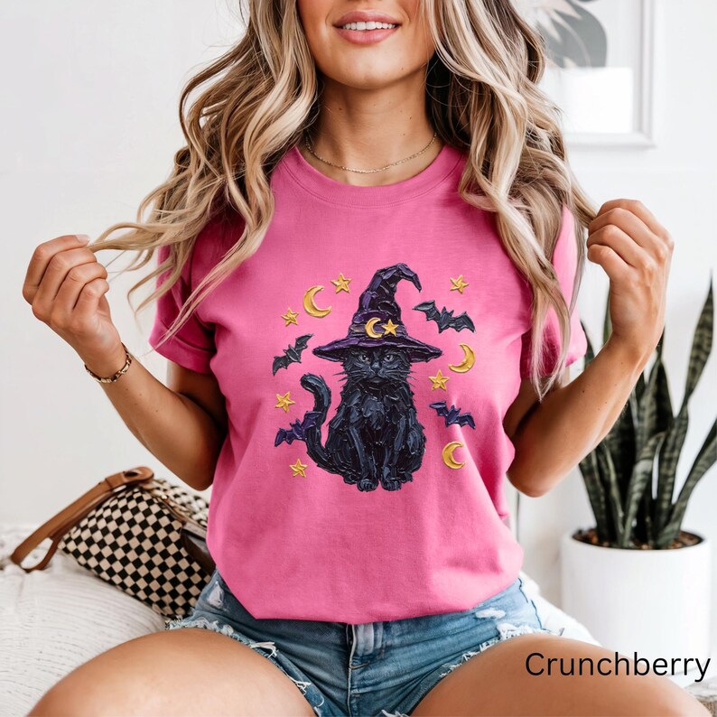 Może przedstawiać: Jaskrawor&oacute;żowy T-shirt z kr&oacute;tkim rękawem z nadrukiem graficznym przedstawiającym czarnego kota w fioletowym kapeluszu czarownicy. Wz&oacute;r zawiera ż&oacute;łte p&oacute;łksiężyce, ż&oacute;łte gwiazdy i ciemnofioletowe nietoperze otaczające kota z przodu koszulki.