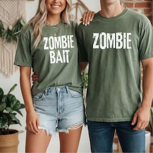 May include: Two olive green short-sleeve t-shirts. One shirt features the white distressed text ZOMBIE BAIT, and the other features the white distressed text ZOMBIE. Both shirts have a crew neckline and a standard fit, made of a solid cotton fabric.