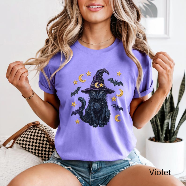 Może przedstawiać: Jasnofioletowy t-shirt z kr&oacute;tkim rękawem z nadrukiem graficznym przedstawiającym czarnego kota w spiczastym kapeluszu czarownicy. Wz&oacute;r zawiera ż&oacute;łte p&oacute;łksiężyce, małe gwiazdki oraz kilka latających nietoperzy otaczających kota z przodu.