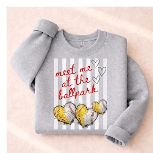 May include: A gray sweatshirt with the text "meet me at the ballpark" in red script. The design includes three baseball-themed hearts and vertical white stripes. The sleeves are cuffed.