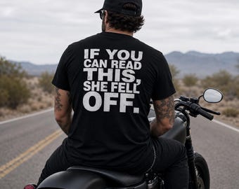 Peut inclure: Un t-shirt noir à manches courtes avec un texte blanc au style vieilli imprimé au dos qui indique : IF YOU CAN READ THIS, SHE FELL OFF. Le vêtement est porté par une personne assise sur une moto sur une route dans un paysage désertique.