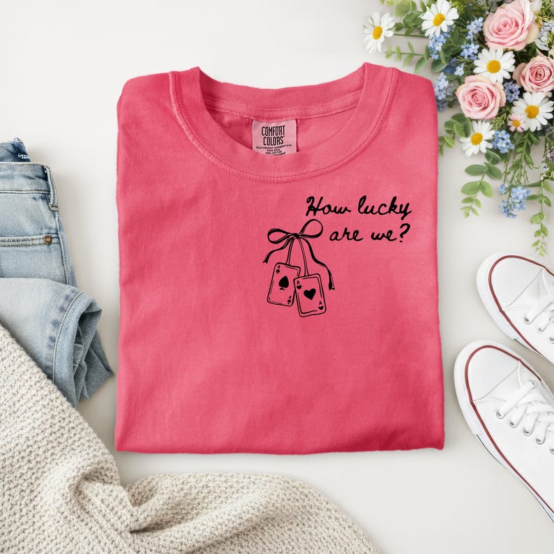 May include: A coral-coloured t-shirt with the text "How lucky are we?" and two playing cards. The shirt is folded, with white trainers and a bouquet of flowers in the background. The shirt has a Comfort Colors tag.