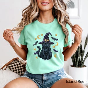 Może przedstawiać: Miętowy t-shirt z kr&oacute;tkim rękawem z nadrukiem graficznym przedstawiającym czarnego kota w fioletowym kapeluszu czarownicy. Wz&oacute;r zawiera ż&oacute;łte p&oacute;łksiężyce, złote gwiazdy i małe fioletowe nietoperze otaczające kota na przodzie koszulki.