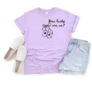 May include: A lilac t-shirt featuring the phrase "How lucky are we?" and a graphic of two playing cards. The shirt is styled with denim shorts and white slip-on shoes.