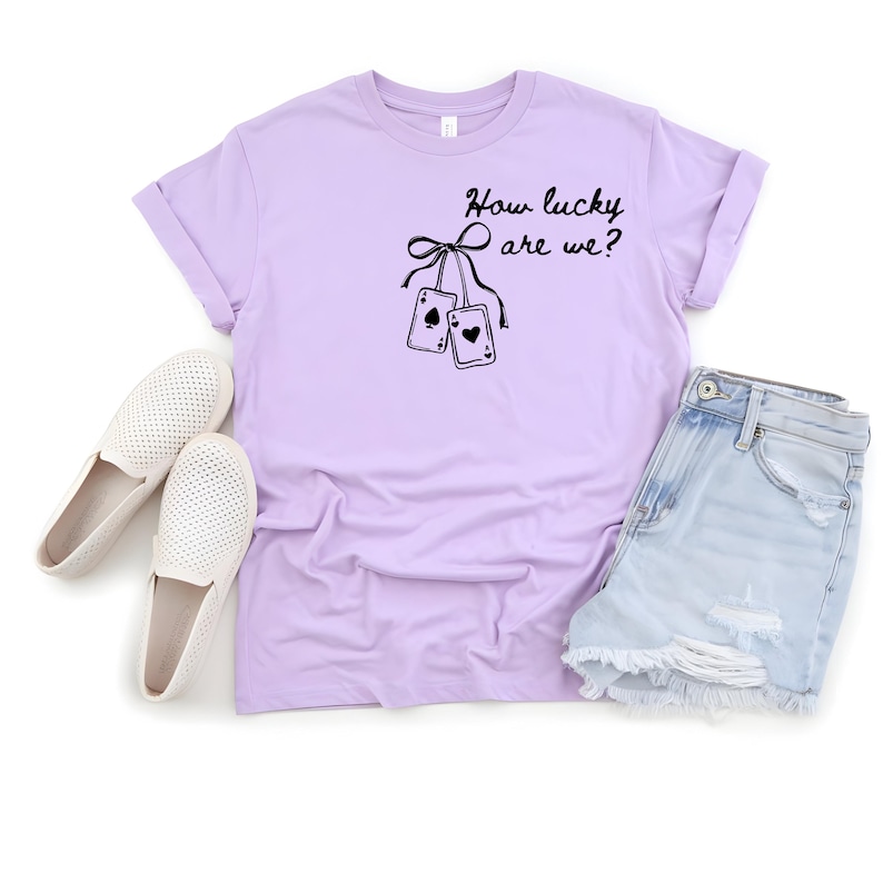 May include: A lilac t-shirt featuring the phrase "How lucky are we?" and a graphic of two playing cards. The shirt is styled with denim shorts and white slip-on shoes.