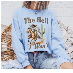 May include: A light blue sweatshirt featuring a graphic of a cowgirl riding a horse, with a cactus and the text "The Hell I Won't." The design is in shades of brown and tan. The sweatshirt is a casual style.