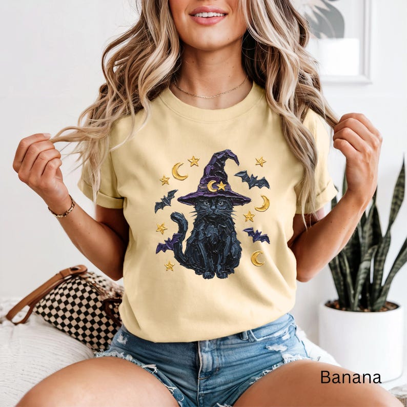 Może przedstawiać: Bladoż&oacute;łty t-shirt z grafiką przedstawiającą czarnego kota w fioletowym kapeluszu czarownicy. Wz&oacute;r zawiera ż&oacute;łte p&oacute;łksiężyce, gwiazdy i małe fioletowe nietoperze otaczające kota. Koszulka ma kr&oacute;tkie rękawy i okrągły dekolt.
