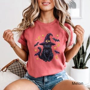 Może przedstawiać: Koralowy t-shirt z kr&oacute;tkim rękawem z nadrukiem czarnego kota w fioletowym kapeluszu czarownicy z ż&oacute;łtym p&oacute;łksiężycem i gwiazdą. Wz&oacute;r zawiera ż&oacute;łte gwiazdy, ż&oacute;łte p&oacute;łksiężyce oraz małe, ciemnoniebieskie latające nietoperze.