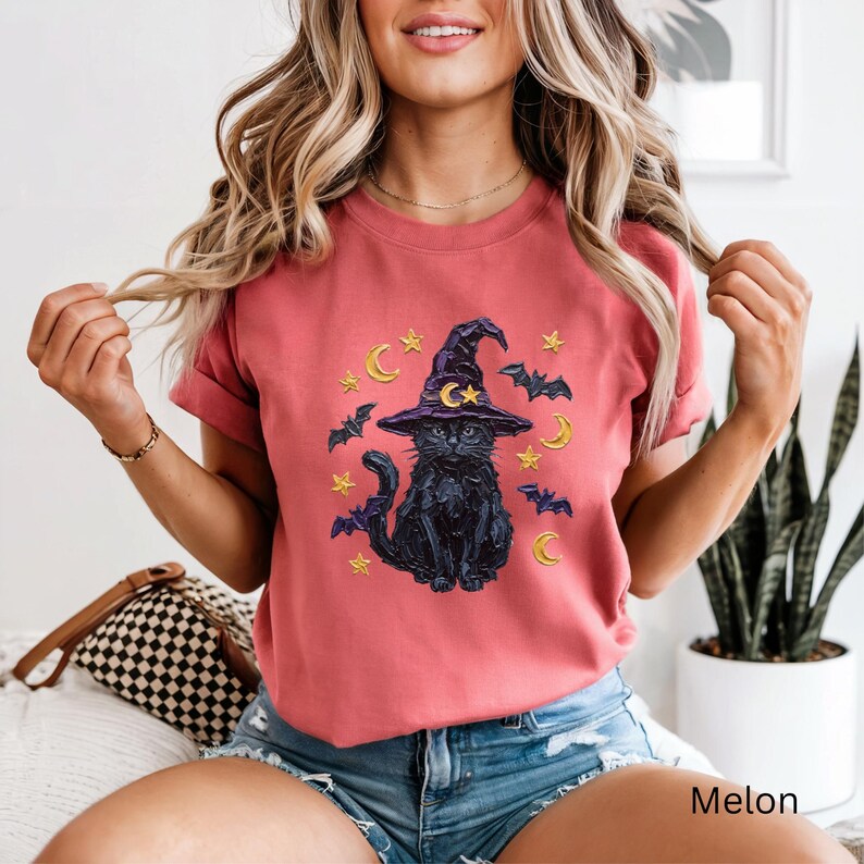 Może przedstawiać: Koralowy t-shirt z kr&oacute;tkim rękawem z nadrukiem czarnego kota w fioletowym kapeluszu czarownicy z ż&oacute;łtym p&oacute;łksiężycem i gwiazdą. Wz&oacute;r zawiera ż&oacute;łte gwiazdy, ż&oacute;łte p&oacute;łksiężyce oraz małe, ciemnoniebieskie latające nietoperze.