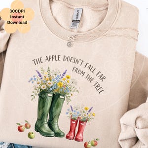 May include: A beige crewneck sweatshirt featuring a graphic of two pairs of rubber boots in green and red, filled with colorful wildflowers. The text reads, The apple doesn't fall far from the tree, accompanied by small red apple illustrations.