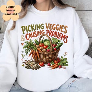 Könnte beinhalten: Weißes Sweatshirt mit einem farbenfrohen Grafikdesign eines Weidenkorbs voller Gemüse, darunter Karotten, Tomaten und Gurken. Der Text "PICKING VEGGIES & CAUSING PROBLEMS" befindet sich über dem Korb. Florale Elemente sind ebenfalls vorhanden.