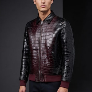 May include: A black and burgundy leather jacket with a crocodile skin texture. The jacket has a zipper closure, a collar, and burgundy ribbed cuffs and hem. The jacket is a bomber style.