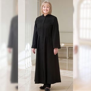 May include: A long, black coat with a high collar and pleated detailing on the chest. The coat has long sleeves and a full-length design. The person is wearing black shoes. The coat appears to be made of a smooth, possibly wool, fabric.