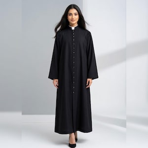 Puede incluir: Una túnica negra larga con cuello blanco y botones frontales. La túnica tiene mangas largas y la lleva una persona. La persona lleva tacones negros. El fondo es una pared blanca lisa.