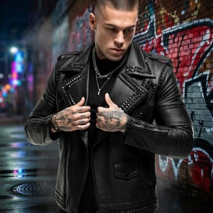 Op de afbeelding: Zwarte leren jas met studs op de kraag, schouders en manchetten. De jas heeft een klassieke biker-stijl met een revers en een ritssluiting aan de voorkant. Het model draagt een zwart shirt en zilveren kettingen.