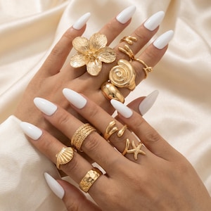 Peut inclure: Une collection de bagues dorées présentant divers motifs, notamment une grande fleur, un coquillage en spirale, une bande texturée, une bande tressée, une étoile de mer et des anneaux ouverts lisses, portées sur plusieurs doigts des deux mains.