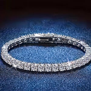 Peut inclure: Un bracelet argenté orné d'une rangée de pierres précieuses rondes et étincelantes. Le bracelet est posé sur une surface bleue, et le fermoir est visible. Les pierres précieuses sont claires et réfléchissent la lumière.