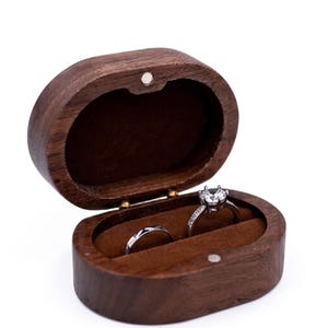 Peut inclure: Une boîte à bagues ovale en bois brun foncé avec un couvercle à charnière et un intérieur doublé de velours brun. À l'intérieur de la boîte, deux bagues argentées sont présentées : un anneau simple et une bague avec une pierre claire.