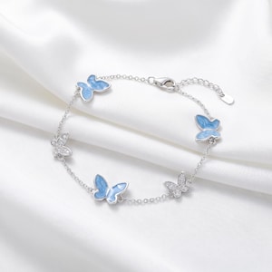 Peut inclure: Un bracelet en chaîne argentée orné de breloques papillon alternées. Deux papillons sont remplis d'émail bleu clair, tandis que les deux autres sont incrustés de petites pierres transparentes. Le bracelet comprend un fermoir mousqueton et une chaînette.