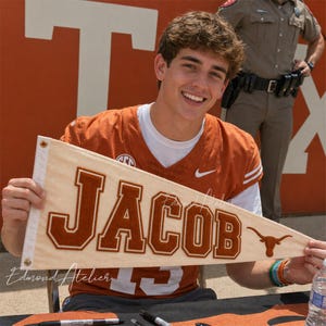 May include: A beige pennant with the name "JACOB" in burnt orange letters and a Longhorn logo. The pennant is held by a person wearing an orange jersey with the Nike logo.