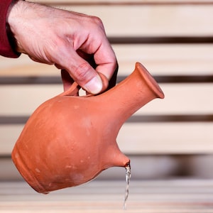 Puede incluir: Una jarra de terracota con asa y pico, sostenida por una persona. El agua se vierte por el pico. La jarra es de color marrón rojizo. El fondo es una superficie de madera de color claro.