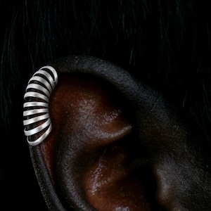 Puede incluir: Un pendiente de oreja plateado con un diseño en espiral se muestra en una oreja de piel oscura sobre un fondo negro. El pendiente de oreja se envuelve alrededor de la parte superior de la oreja, creando un aspecto moderno y minimalista. La joya es de metal brillante.