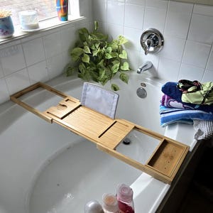 Puede incluir: Una bandeja de bambú para bañera se extiende sobre una bañera blanca. La bandeja tiene compartimentos y un soporte para libros. Botellas de productos de baño están en el borde de la bañera. También se ven una planta y toallas.