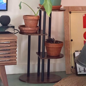 Może przedstawiać: Wielopoziomowy stojak na rośliny z ciemnobrązową podstawą i półkami, podtrzymujący kilka roślin doniczkowych. Stojak ma czarne metalowe podpory i okrągłą podstawę. Doniczki są terakotowe i turkusowe, zawierające różne zielone rośliny. Stojak ma około 122 cm wysokości.