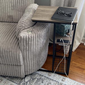 May include: A light gray corduroy recliner chair with a side table. The table has a wood-look top and a black metal frame. A laptop and glasses rest on the table. A patterned rug is on the floor.