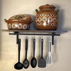 May include: A kitchen shelf with two brown ceramic pots with lids, decorated with white dots. Below, a row of black kitchen utensils hangs, including a ladle, slotted spoon, solid spoon, spatula, and whisk. The shelf is mounted on a gray wall.