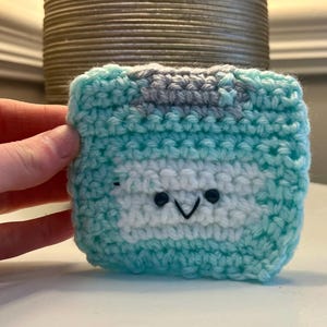 May include: A crocheted, square-shaped case in shades of blue, white, and gray. The case features a cartoon face with black eyes and a smiling mouth. The item is handmade and appears to be a protective cover for a small electronic device.