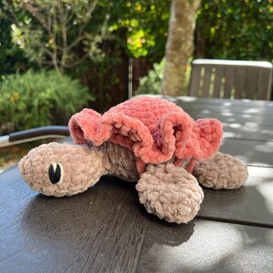 K&ouml;nnte beinhalten: Ein geh&auml;keltes Schildkr&ouml;ten-Stofftier mit einem weichen, hellbraunen K&ouml;rper in flauschiger Textur und einem auff&auml;lligen, ger&uuml;schten Panzer in Altrosa. Die Schildkr&ouml;te hat einfache, aufgestickte schwarz-wei&szlig;e Augen auf ihrem runden Kopf.