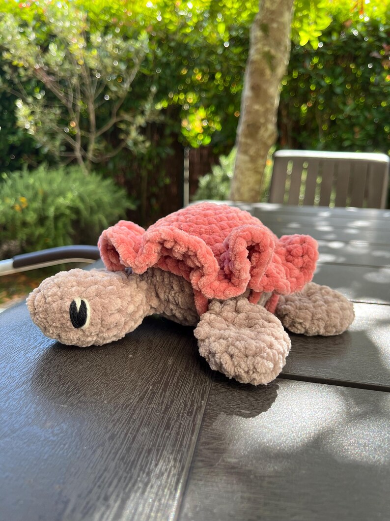K&ouml;nnte beinhalten: Ein geh&auml;keltes Schildkr&ouml;ten-Stofftier mit einem weichen, hellbraunen K&ouml;rper in flauschiger Textur und einem auff&auml;lligen, ger&uuml;schten Panzer in Altrosa. Die Schildkr&ouml;te hat einfache, aufgestickte schwarz-wei&szlig;e Augen auf ihrem runden Kopf.