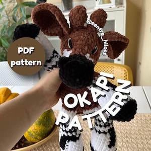 Puede incluir: Un peluche de okapi hecho de hilo marrón, negro y blanco con una textura suave y difusa. El okapi presenta orejas grandes, un hocico oscuro y patas a rayas. El texto en la imagen dice PDF pattern y OKAPI PATTERN con @eentre_hiloss.