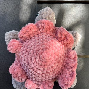 K&ouml;nnte beinhalten: Ein handgefertigtes H&auml;kel-Pl&uuml;schtier in Form einer Schildkr&ouml;te. Der Panzer besteht aus weichem, strukturiertem rosa Garn mit kreisf&ouml;rmigem Muster, w&auml;hrend der Kopf und die vier Flossen aus kontrastierendem hellgrauem Garn mit Pl&uuml;schstruktur gefertigt sind.