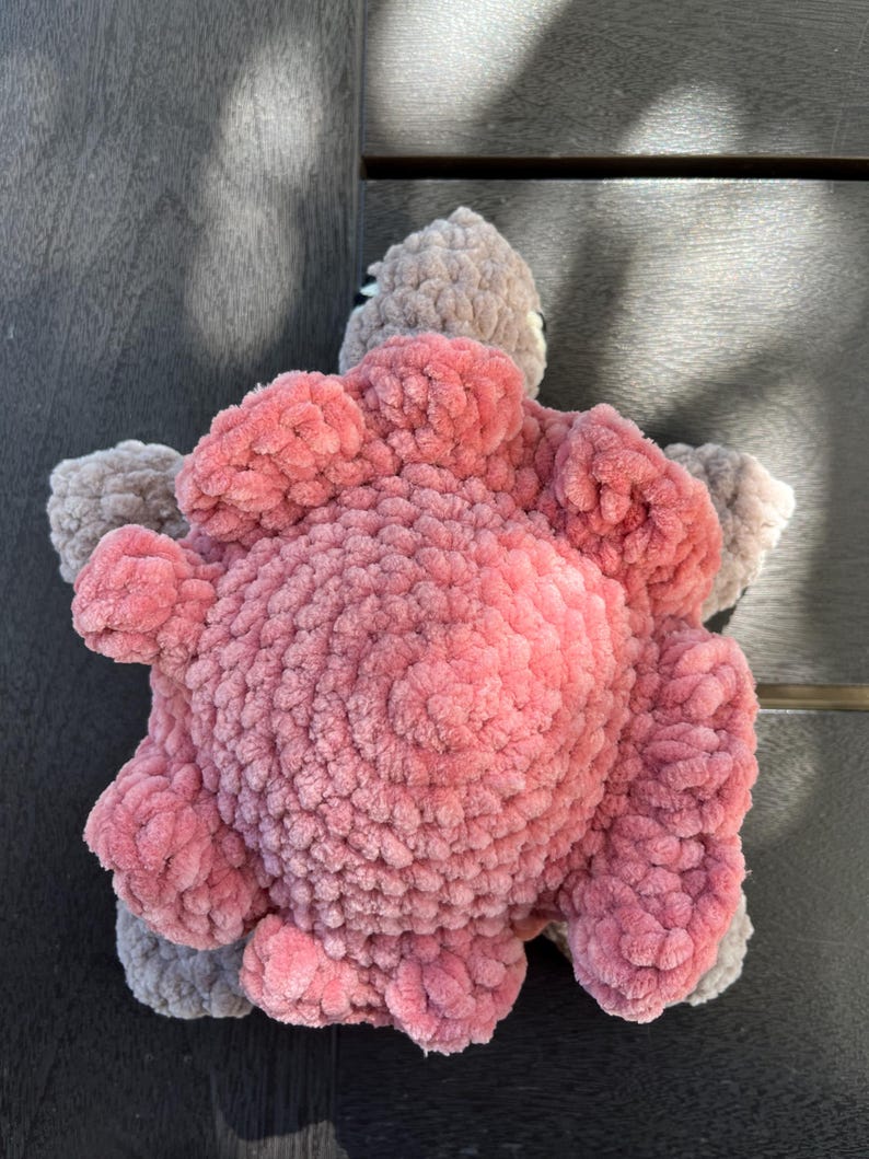 K&ouml;nnte beinhalten: Ein handgefertigtes H&auml;kel-Pl&uuml;schtier in Form einer Schildkr&ouml;te. Der Panzer besteht aus weichem, strukturiertem rosa Garn mit kreisf&ouml;rmigem Muster, w&auml;hrend der Kopf und die vier Flossen aus kontrastierendem hellgrauem Garn mit Pl&uuml;schstruktur gefertigt sind.