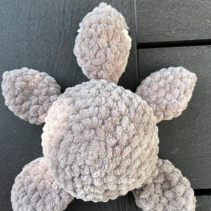 K&ouml;nnte beinhalten: Ein Pl&uuml;sch-Schildkr&ouml;tenspielzeug aus weichem, hellbraunem Chenille-Garn. Das Stofftier verf&uuml;gt &uuml;ber einen abgerundeten zentralen Panzer, einen ausgepr&auml;gten Kopf mit sichtbaren wei&szlig;en Augendetails und vier abgerundete Flossen am K&ouml;rper.