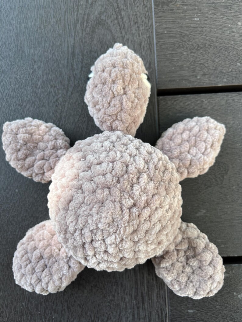 K&ouml;nnte beinhalten: Ein Pl&uuml;sch-Schildkr&ouml;tenspielzeug aus weichem, hellbraunem Chenille-Garn. Das Stofftier verf&uuml;gt &uuml;ber einen abgerundeten zentralen Panzer, einen ausgepr&auml;gten Kopf mit sichtbaren wei&szlig;en Augendetails und vier abgerundete Flossen am K&ouml;rper.