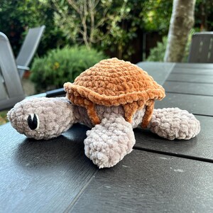 K&ouml;nnte beinhalten: Ein Pl&uuml;schschildkr&ouml;ten-Spielzeug aus weichem, strukturiertem Garn. Die Schildkr&ouml;te hat einen hellgrauen Kopf, Gliedma&szlig;en und einen Bauch, die mit einem abgerundeten, dunkelbraunen Panzer kontrastieren. Sie hat einfache schwarze Augen.
