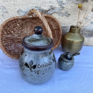 Puede incluir: Un tarro de cerámica con tapa, etiquetado "Cerises", y un pequeño tarro a juego con una cuchara. Los tarros son grises con detalles marrones y un diseño de hojas. Una cesta de mimbre y un jarrón dorado están en el fondo.