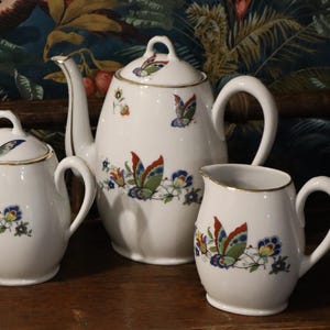 Op de afbeelding: Een wit porseleinen theeservies met een theepot, suikerpot en melkkan. Elk stuk is versierd met kleurrijke vlinder- en bloemmotieven. De set heeft gouden randen en staat op een houten oppervlak.