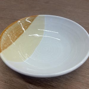 May include: A ceramic bowl with a distinctive design. The bowl has a white interior and a section of tan and cream-coloured glaze on the rim. The bowl is round and has a simple, elegant design.