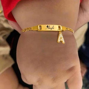 May include: A gold-colored bracelet with the name "Angle" engraved on a rectangular plate. A charm in the shape of the letter "A" is attached. The bracelet is on a wrist.