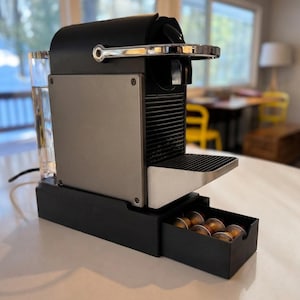 Pode incluir: Uma máquina de café expresso preta e prateada com uma gaveta retrátil contendo cápsulas de café. Um copo de água está à esquerda. A máquina está em uma bancada branca.