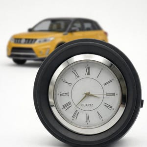 Op de afbeelding: Een zwarte en zilveren quartz klok met Romeinse cijfers en een zilveren rand. De klok staat tegen een vage achtergrond met een gele auto.