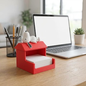 Op de afbeelding: Een rode Snoopy-thema notitieblokhouder met een witte Snoopy-figuur erop. De houder heeft de vorm van een hondenhok en bevat een stapel wit notitiepapier. Een laptop, pennen en een kleine plant staan op de achtergrond.