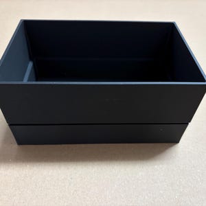 Könnte beinhalten: Eine schwarze rechteckige Box mit zweistufigem Design. Der obere Bereich ist offen und zeigt einen leeren Innenraum. Die Box besteht aus einem mattschwarzen Material und steht auf einer hellbraunen Oberfläche.