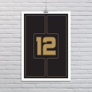 May include: A framed poster with a black background and the number 12 in gold. The number is centered and outlined in gold, with a gold border around the black background. The poster is hanging on a white brick wall.