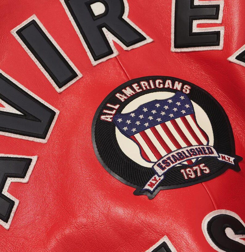 May include: Close-up of a red leather jacket with black and white lettering spelling "AIR FORCE". A circular patch features the American flag and the words "ALL AMERICANS ESTABLISHED 1975 N.Y."