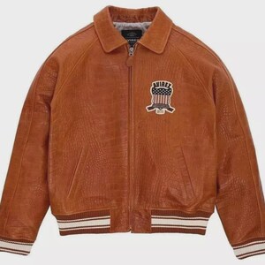 Puede incluir: Chaqueta de cuero marr&oacute;n con acabado texturizado, cuello y cierre de cremallera. La chaqueta tiene un parche en el pecho con un dise&ntilde;o de bandera americana y la palabra "AVIREX". Los pu&ntilde;os y el dobladillo tienen un dise&ntilde;o a rayas.
